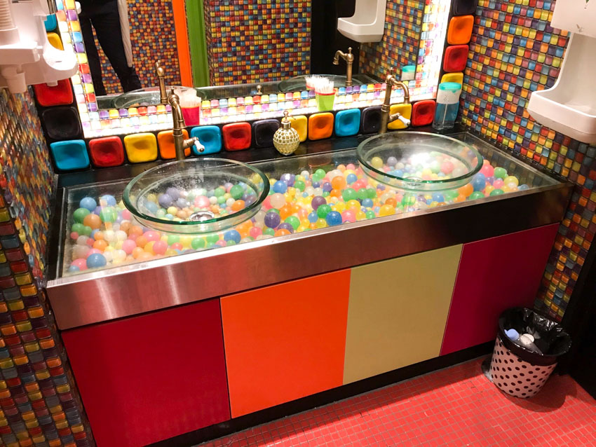 Two basins in front of a mirror, bordered by giant colourful tiles. The basins are transparent, revealing colourful balls sitting in a bed underneath.