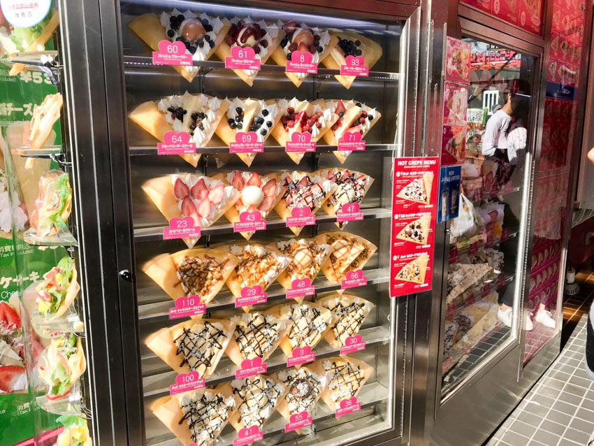 Many fake plastic crepes on display in a glass store front, indicating various flavours
