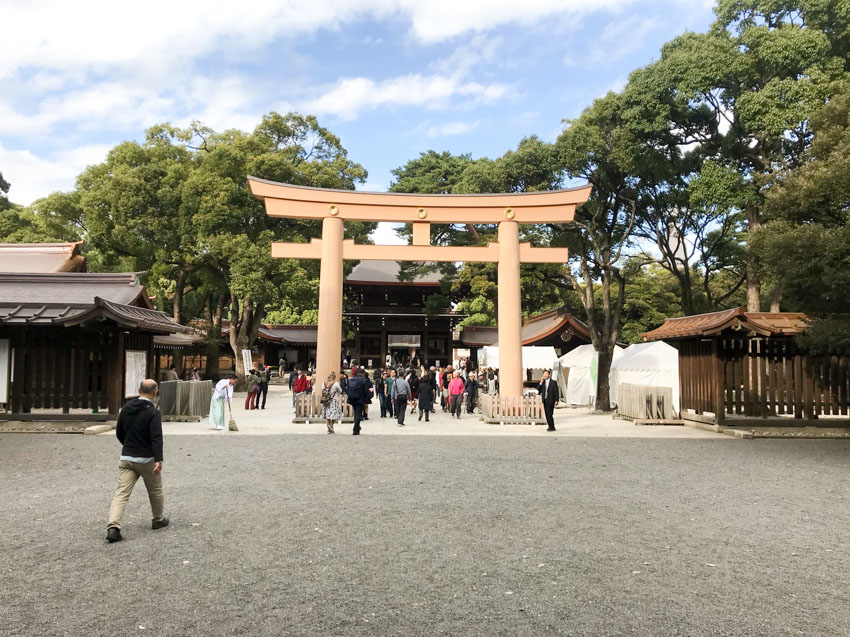 Large gates at the entrance to a shrine. Some low buildings and big green trees are within the grounds to the shrine. A man walks towards the entrance from the left, but close in proximity