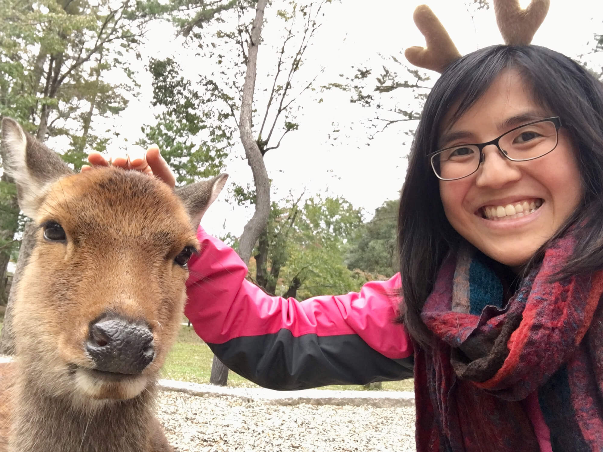 A woman with glasses, smiling and patting a deer