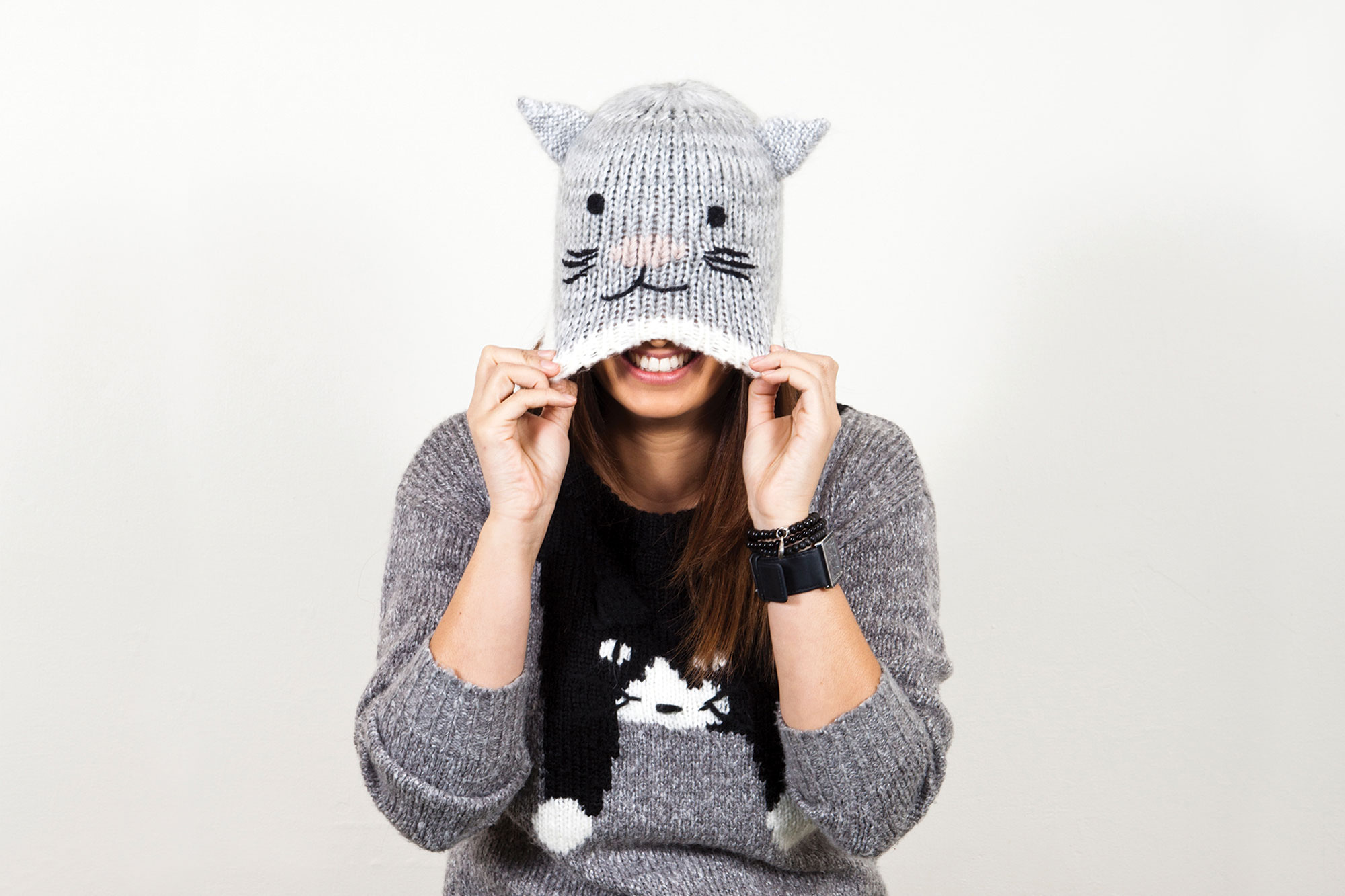Kylie, wearing a grey sweater with a cat image down the front, and a light grey cat shaped beanie pulled over her eyes with her hands. She is grinning.