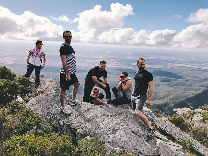Six people atop a rocky mountain, in various different poses. The sky is blue and has quite a lot of white clouds.