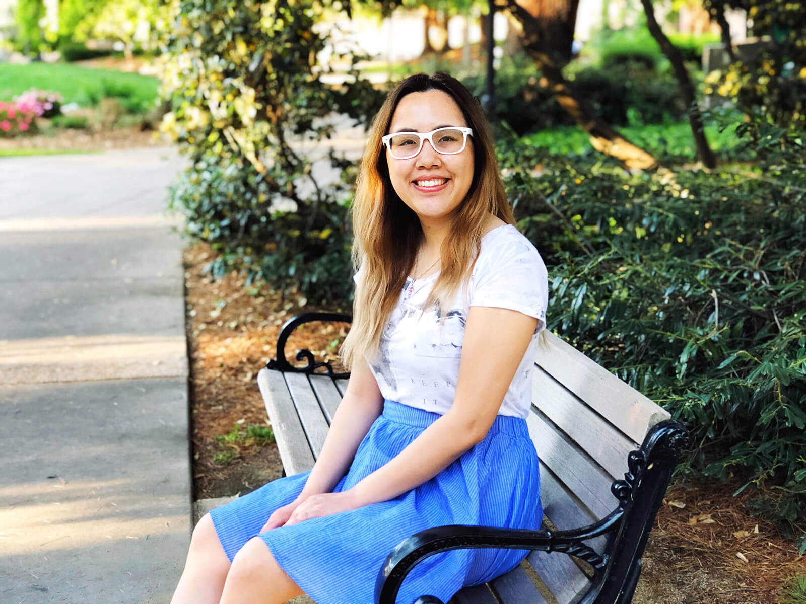 A girl sitting on a park bench, smiling. She has dark hair with blonde highlights, is wearing white glasses, and a light coloured top and a blue skirt.