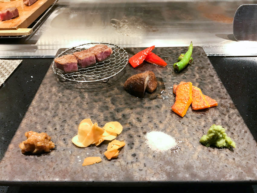 A square stone plate with some vegetables and kobe beef cooked and served, along with some condiments