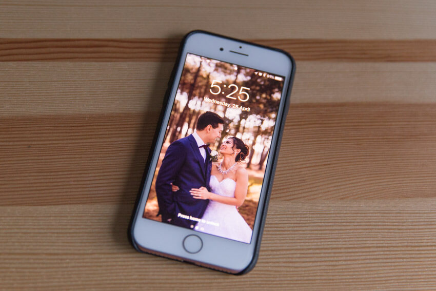An iPhone with a black leather case, screen-up on a wooden surface, showing a man and woman in traditional wedding clothing on the screen