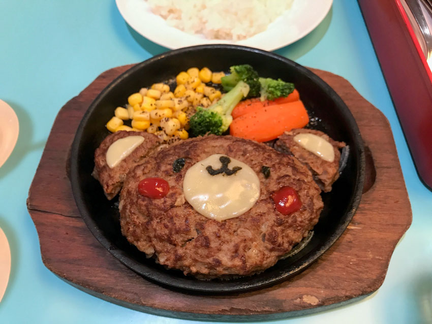 A hamburger steak shaped in an oval with a bear’s face created on it with melted cheese, tomato sauce and tiny pieces of seaweed. Two small semicircles of beef patty with melted cheese on top form ears for the bear’s face. The meal is served on a hot plate with carrot, corn and broccoli.