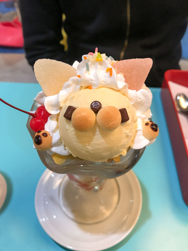 Birds-eye view of a parfait dessert in a tall glass with squiggly edges. The contents of the parfait are shaped like a cat, with a scoop of ice cream forming the face, small ball-shaped cookies forming the hands and mouth/nose, cylindrical chocolate pellets forming the eyes, and sugar-coated jellies forming the ears. The dessert is adorned with white whipped cream, colourful sprinkles and a bright red cherry.