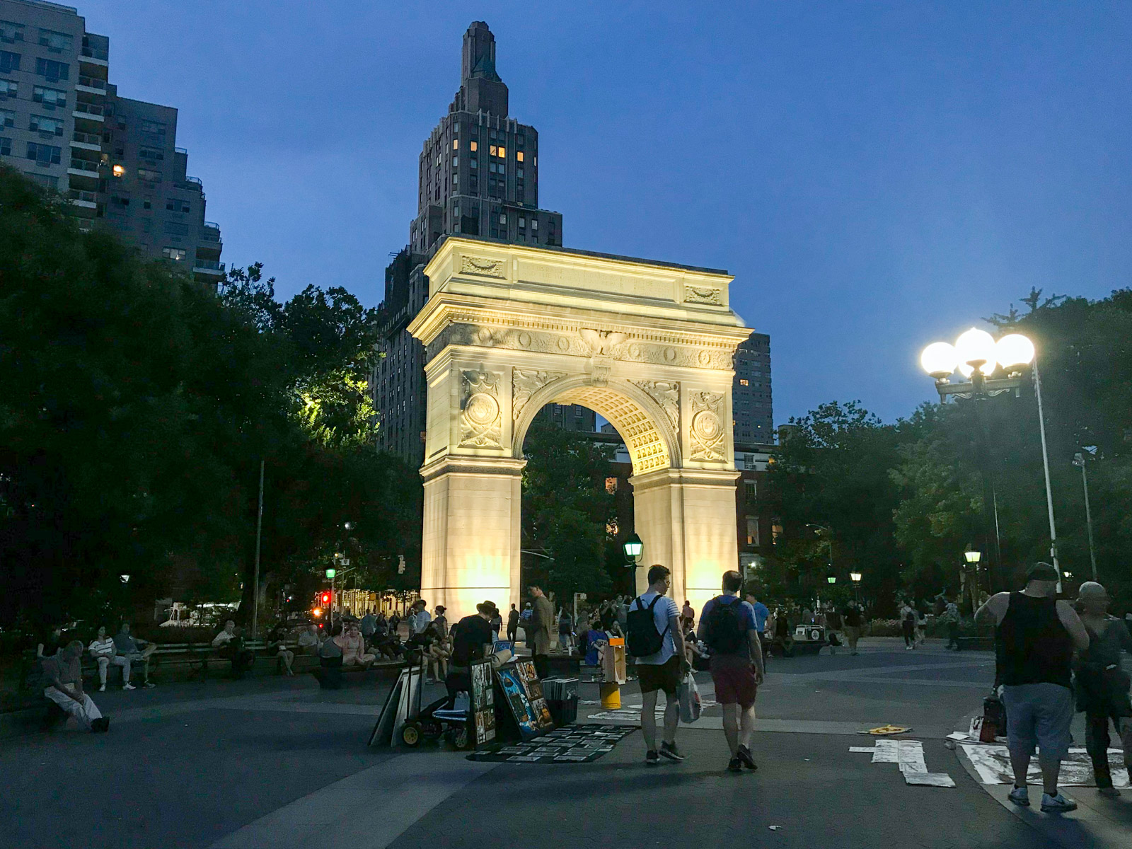 Washington Square Arch, a tall arch structure, lit up in the evening. In the foreground, people are sharing their artwork with it laid out on the ground