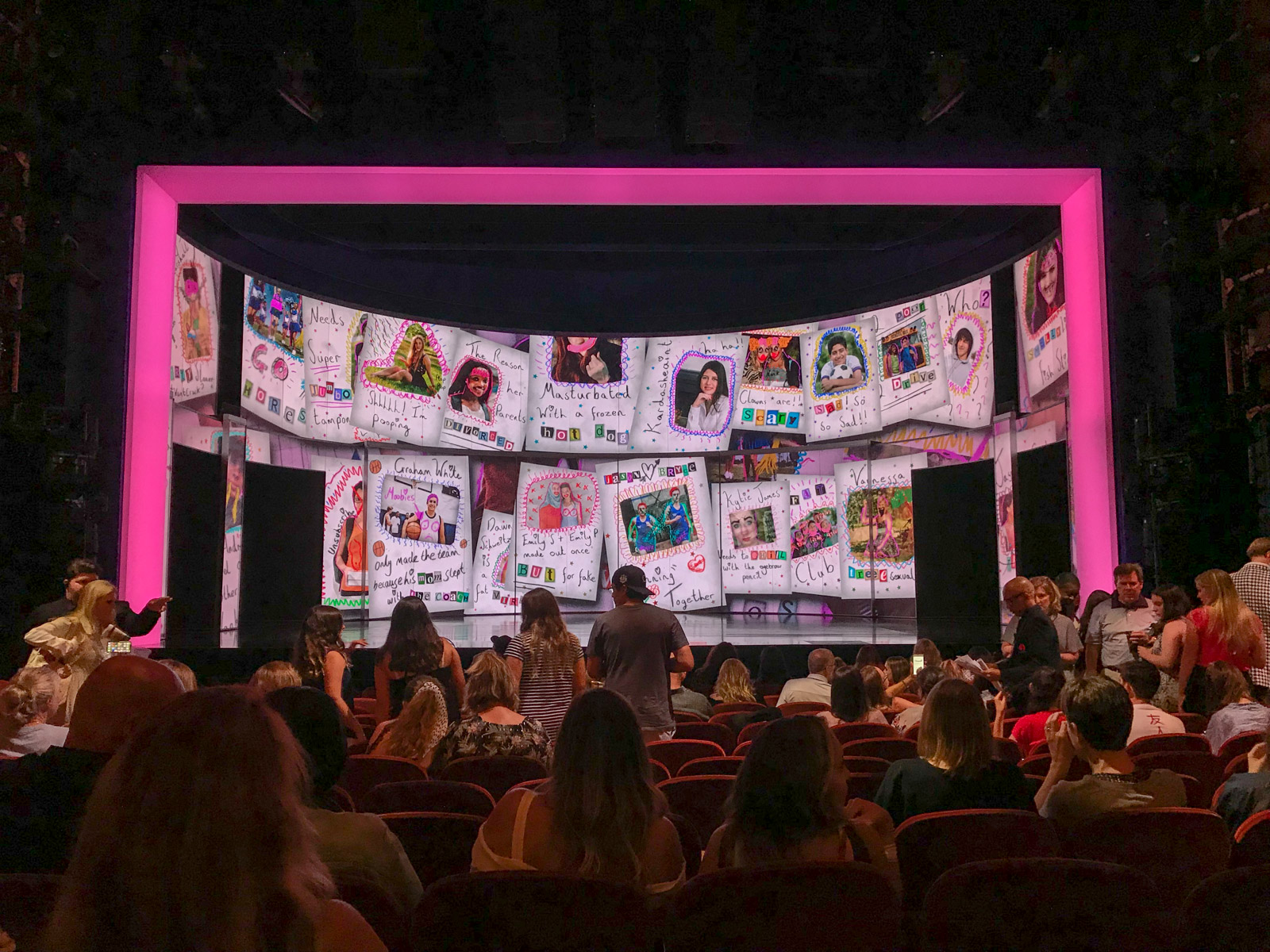 The inside of a theatre with audience members both sitting and making their way to their seats. The stage is ready with a digital backdrop resembling a scrapbook of photos and comments