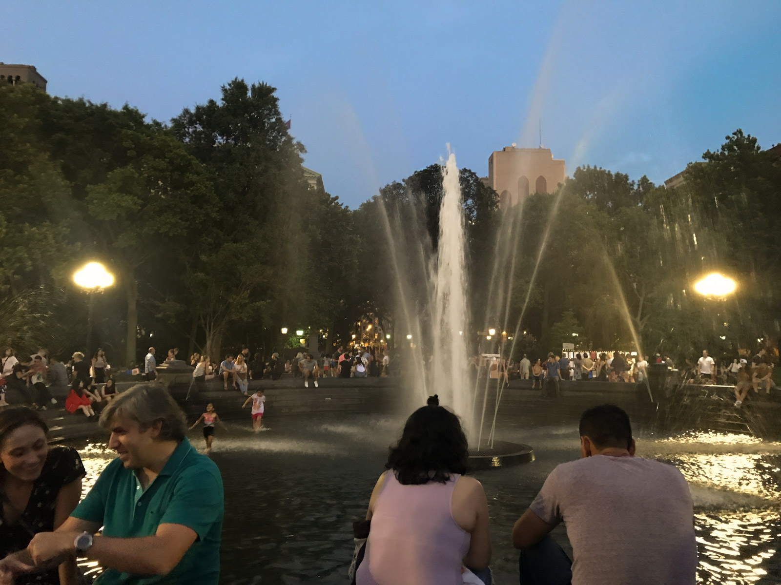 A nighttime view of many people sitting around a fountain. The fountain is active and jetting water.