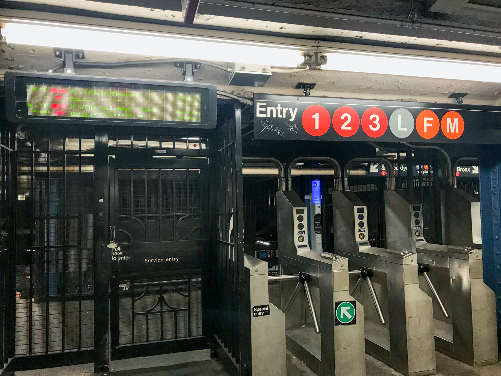 A row of turnstiles with subway lines printed on a sign above them. To the left is a black service entry/exit gate that resembles a door