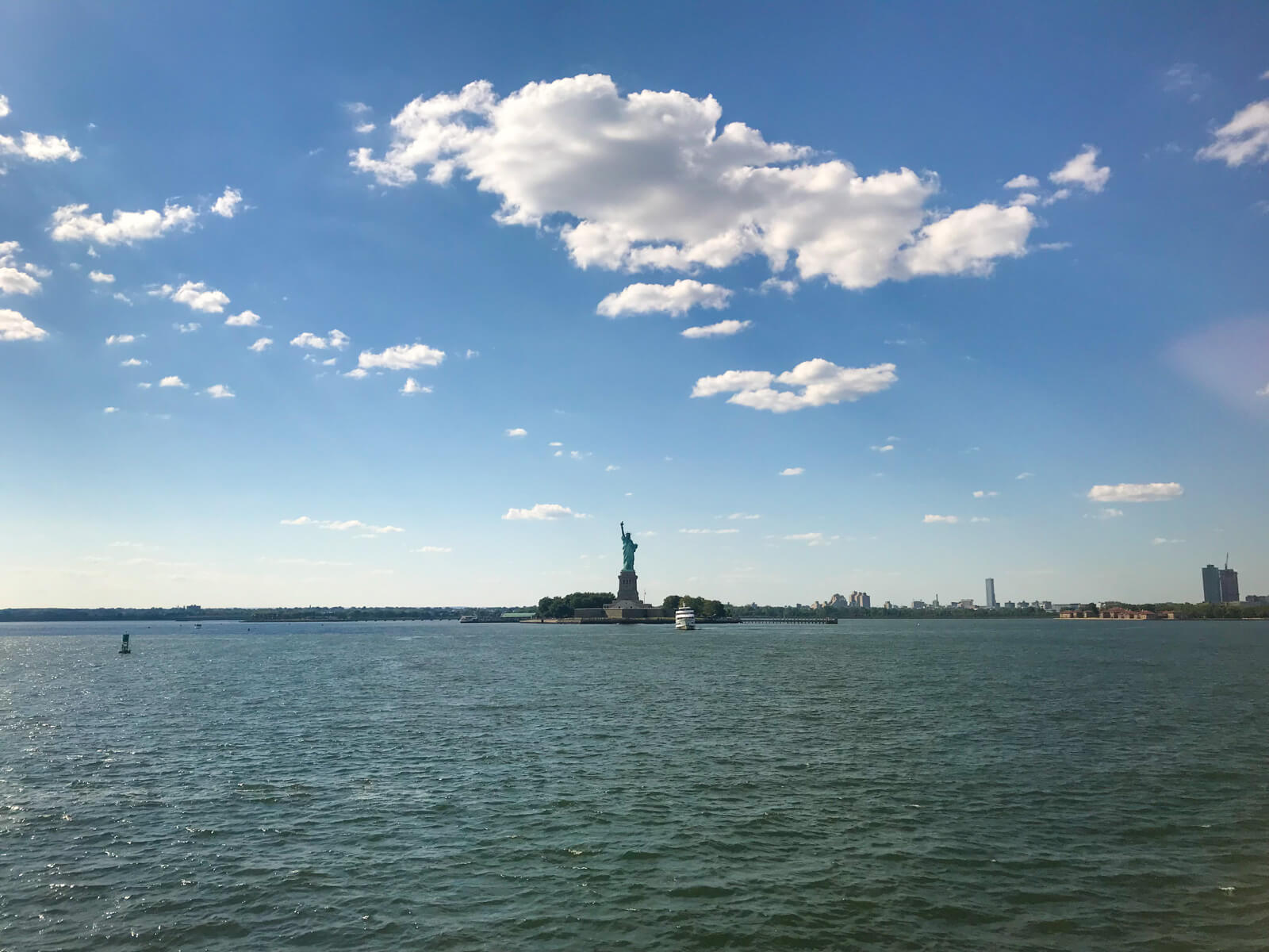 A view from a ferry of the Statue of Liberty, with a lot of the ocean visible around it. The sky is blue and has few clouds