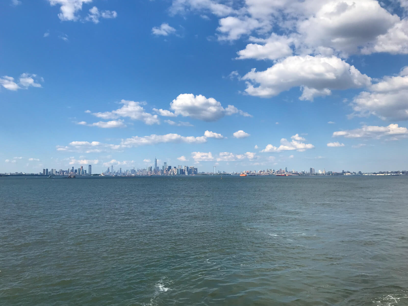 A city skyline with the ocean taking up the bottom half of the frame and blue sky with many clouds taking up the top half of the frame