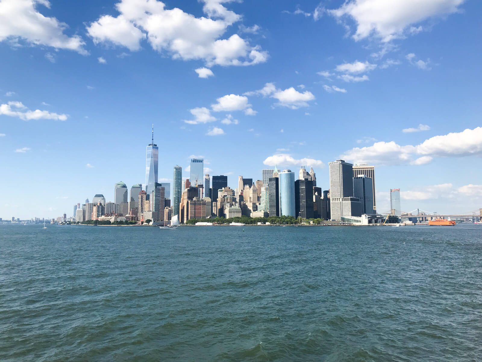 A view of the New York City skyline with the ocean in the foreground. The sky is blue with several clouds
