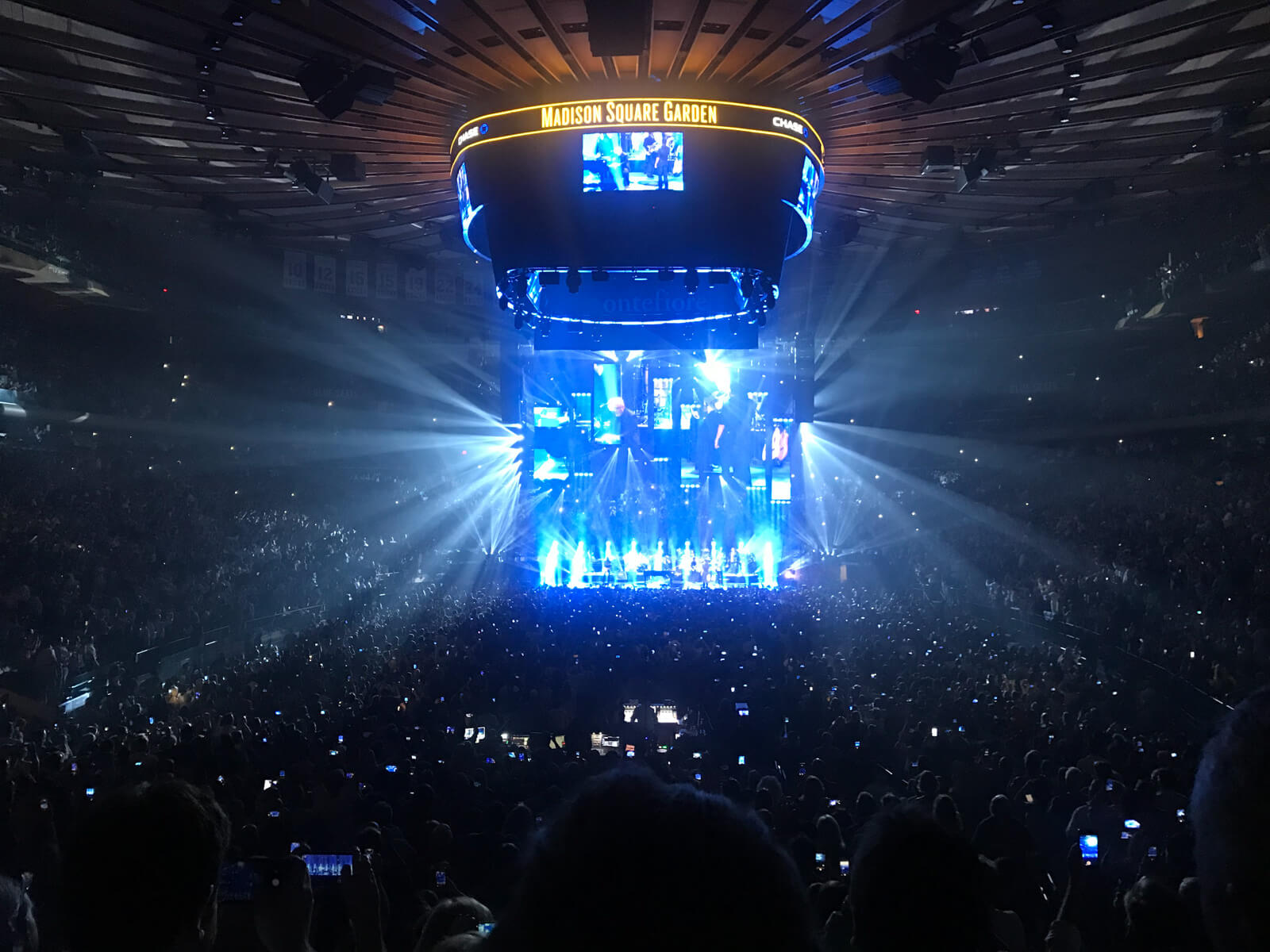 The inside of a concert venue. The stage has a lot of blue lighting and a fixture above the stage and on the ceiling of the venue reads “Madison Square Garden” and has screens with close views of what is happening on stage