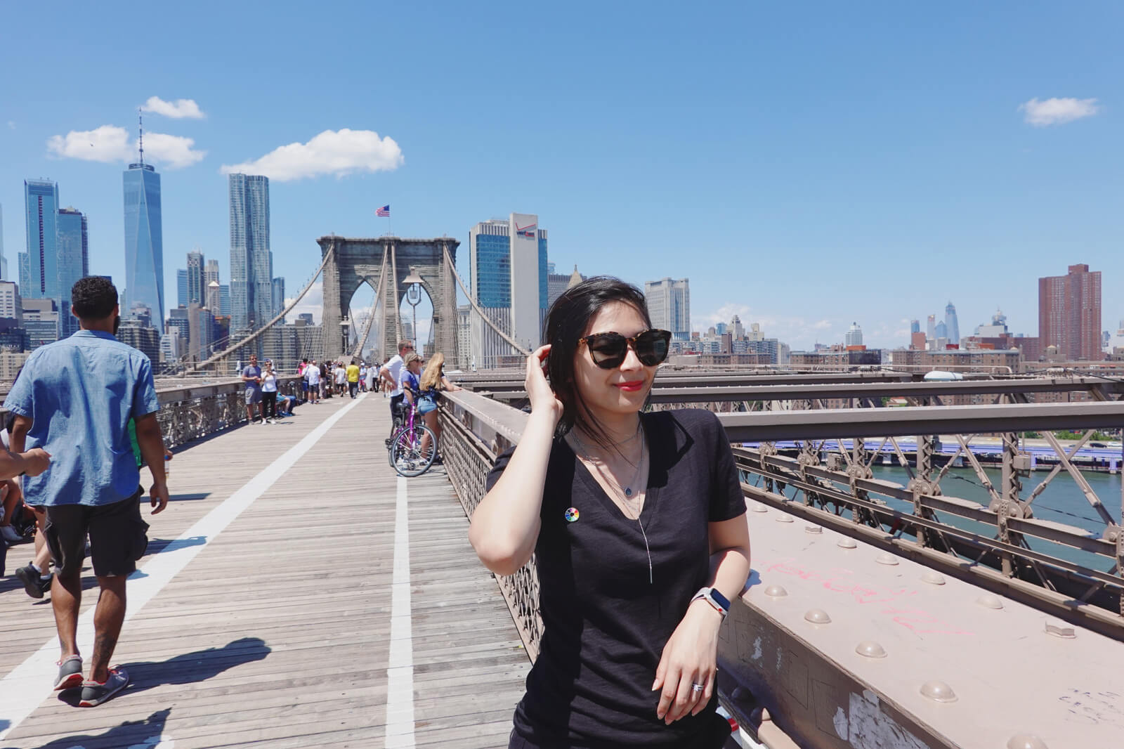 A woman in a black t-shirt, wearing sunglasses. She is standing on a bridge with high-rise buildings in the background. There are other people bicycling and standing on the bridge