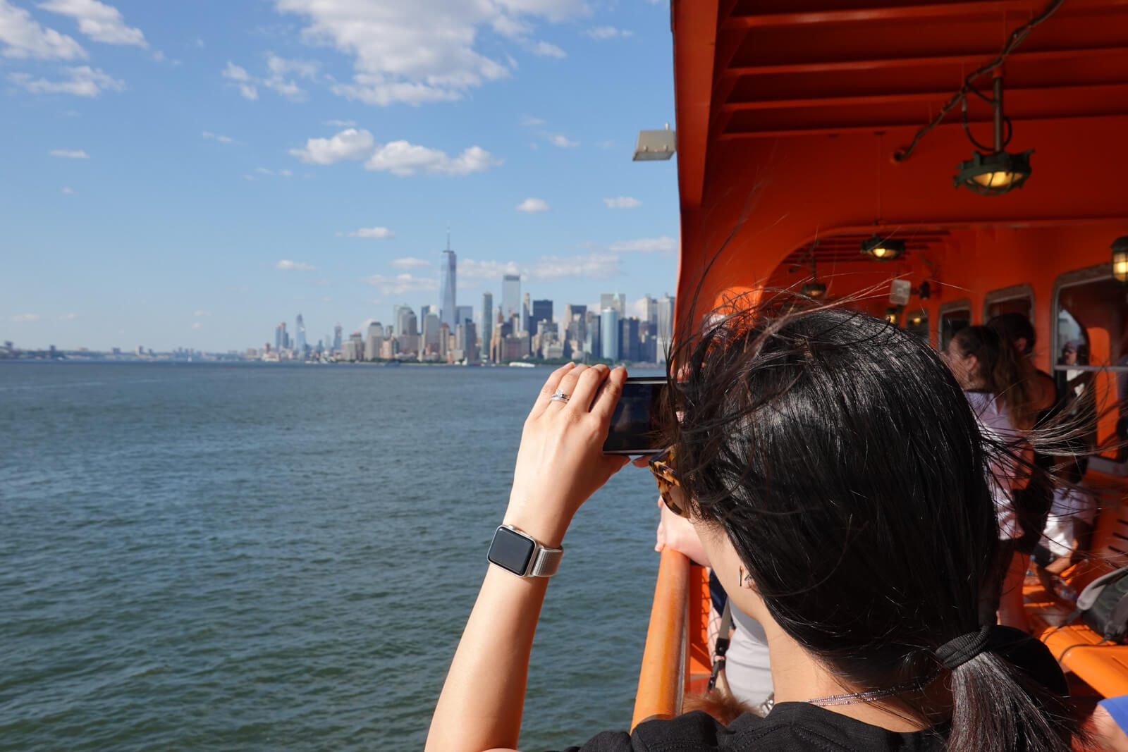 The back of a woman’s head as she takes a photo with her smartphone from a ferry. In the distance is a city with high-rise buildings