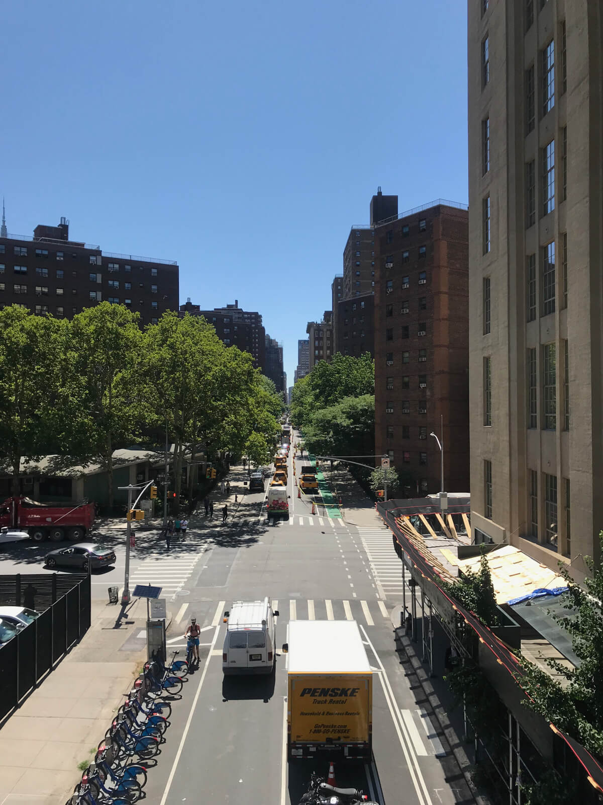 A daytime, high-angle view of a street in New York on a sunny day. Many bicycles are parked on the side of the street and there are a couple of vans and small trucks driving on the street