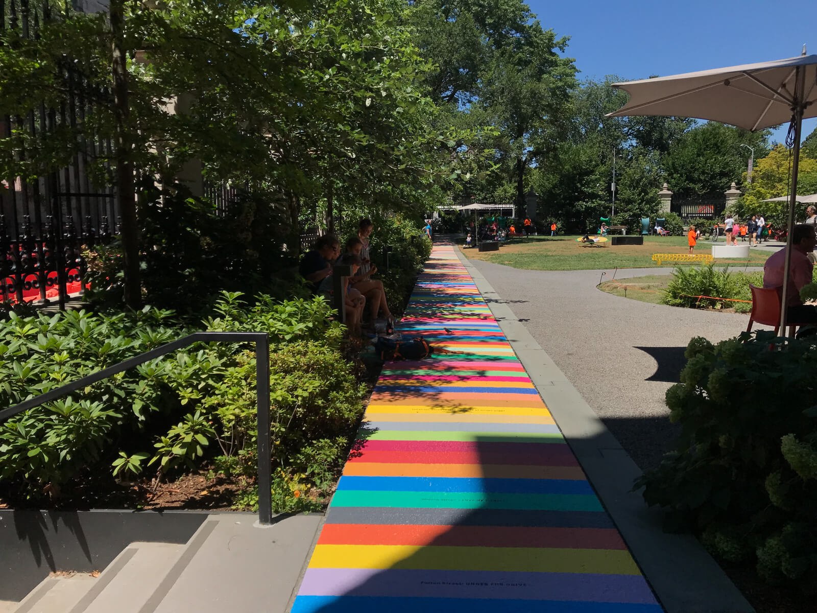 The inside of a park with a horizontally-striped short path in different colours. Some trees provide shade on the path