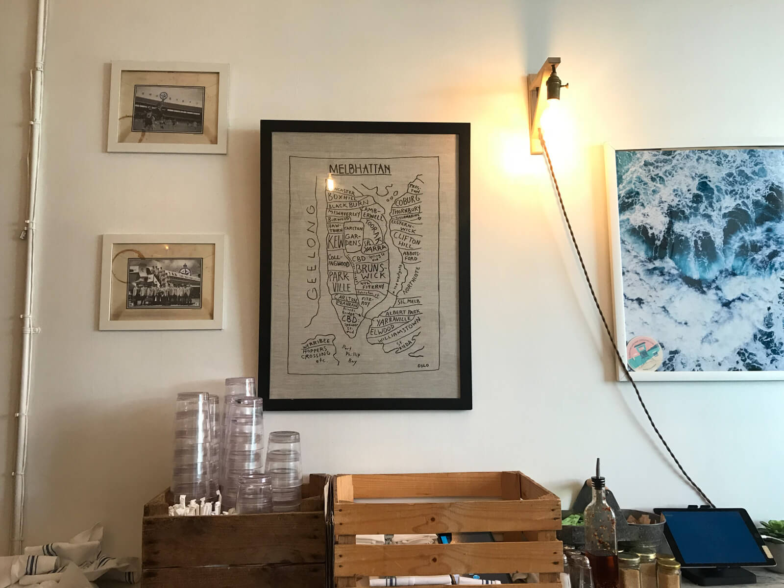 The interior decor of a cafe, with black-and-white photographs on the wall and a piece of drawn artwork titled “Melbhattan”, with outlines and labels resembling a map of a fictitious area