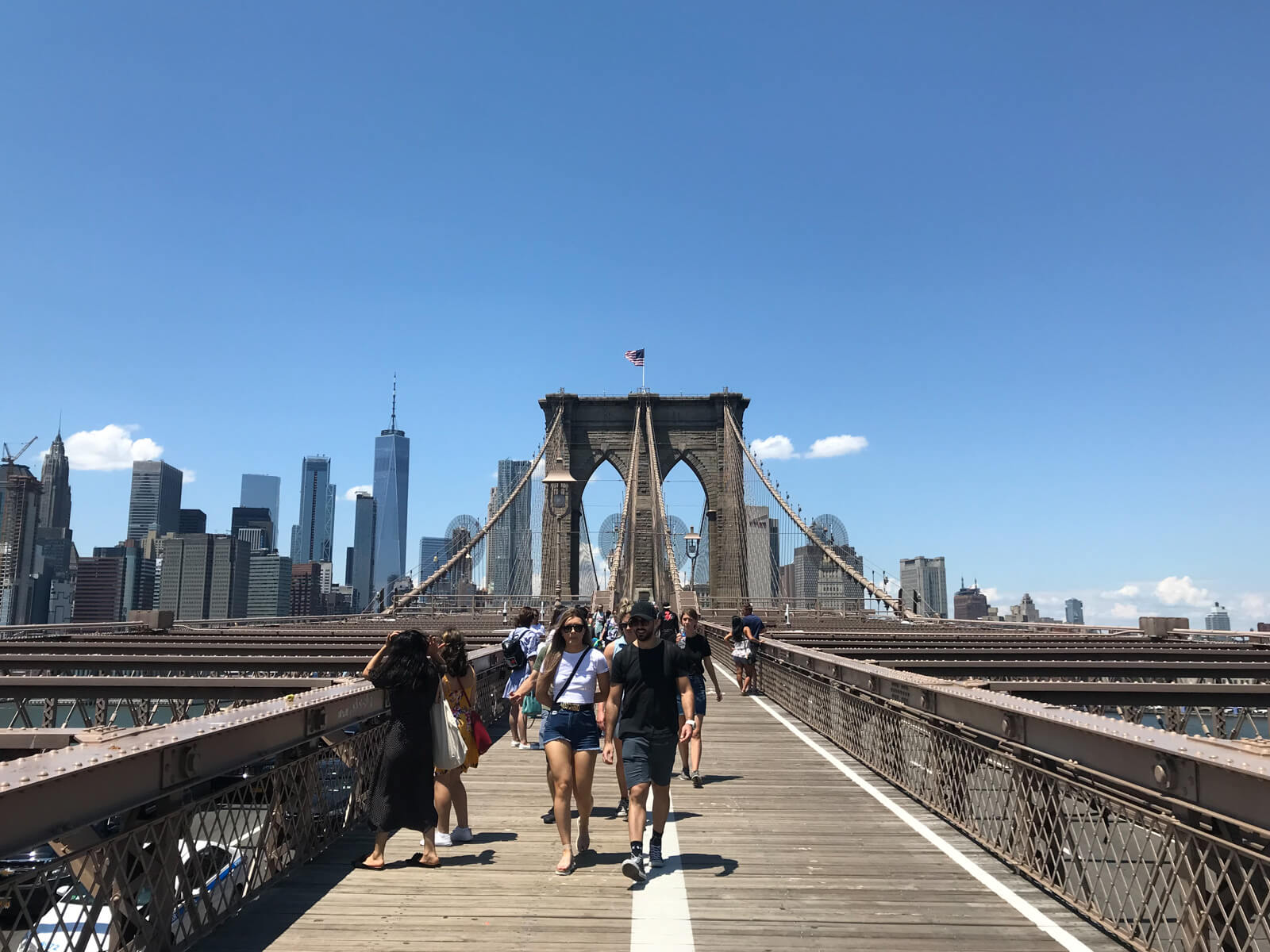 The Brooklyn Bridge as seen from the view of someone walking on it. There are some people on the bridge walking and others standing to the sides