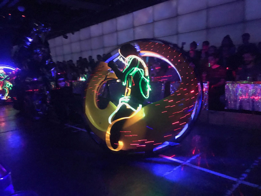 In a dark room with an open area, a person sits on a bike that is the shape of a large circular wheel. The person is in a dark costume with glowing wires woven throughout it. Shadows of an audience can be seen in the background.