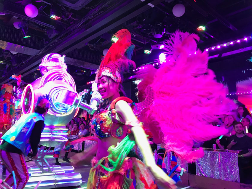 A girl dressed up in a costume consisting of a crop top and skirt, with big feather wings attached to her back. Purple lighting from the ceiling of the room colours her face and wings. In the background is a tall metallic robot.