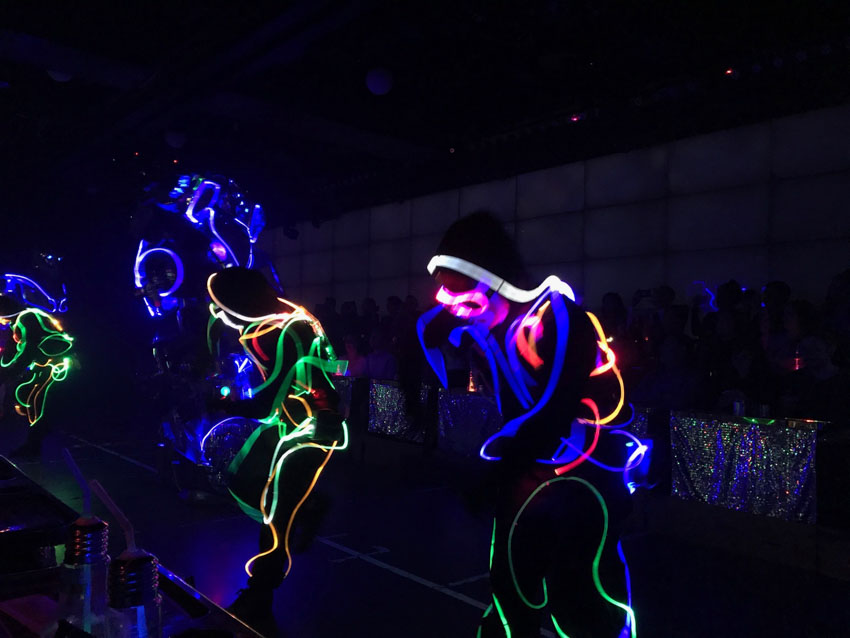 A dark room with several people in it, mid-dance, wearing black costumes with glowing wires woven through.