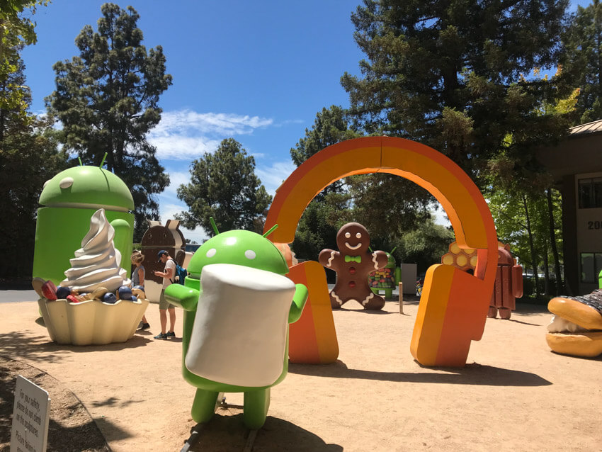 A dirt ground with various sculptures: a giant orange set of headphones; a frozen yoghurt with berries; the Google Android robot holding a marshmallow; as well as some others in the background