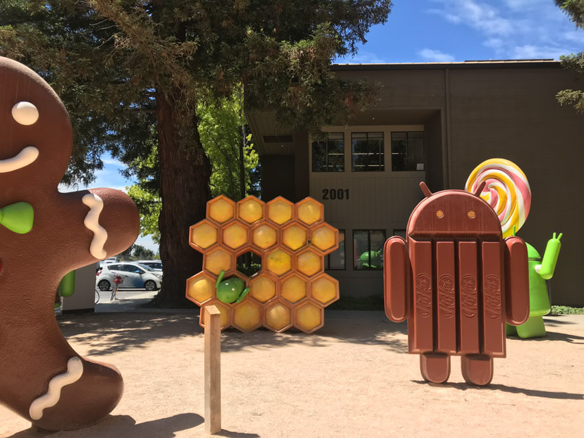 A dirt ground with various sculptures, a couple of metres tall. One resembles a robot made of chocolate Kit Kats, another resembles honeycomb, and another two can be partially seen: a green robot holding a lollipop, and a giant gingerbread man