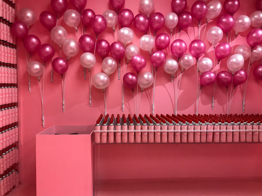 A pink painted room with a table full of pink aerosol cans. There are pink balloons attached to the wall.