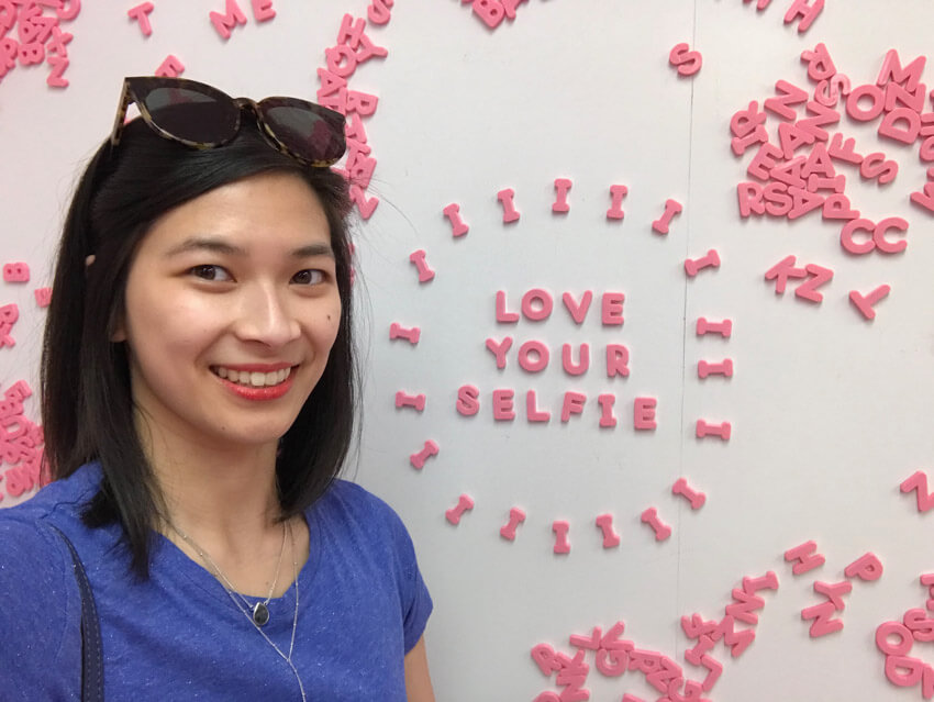 A woman with short dark hair in a blue shirt, smiling. Behind her is a whiteboard reading “love your selfie” in pink magnetic letters