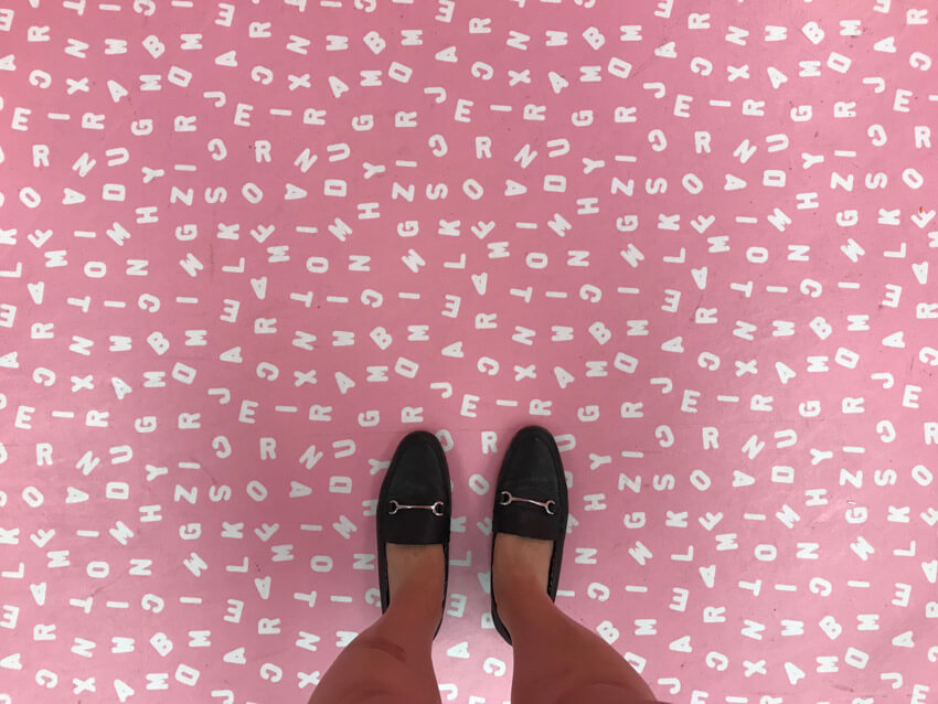 A light pink floor with a pattern of white uppercase letters. A woman’s feet wearing black loafers are at the edge of the photo.