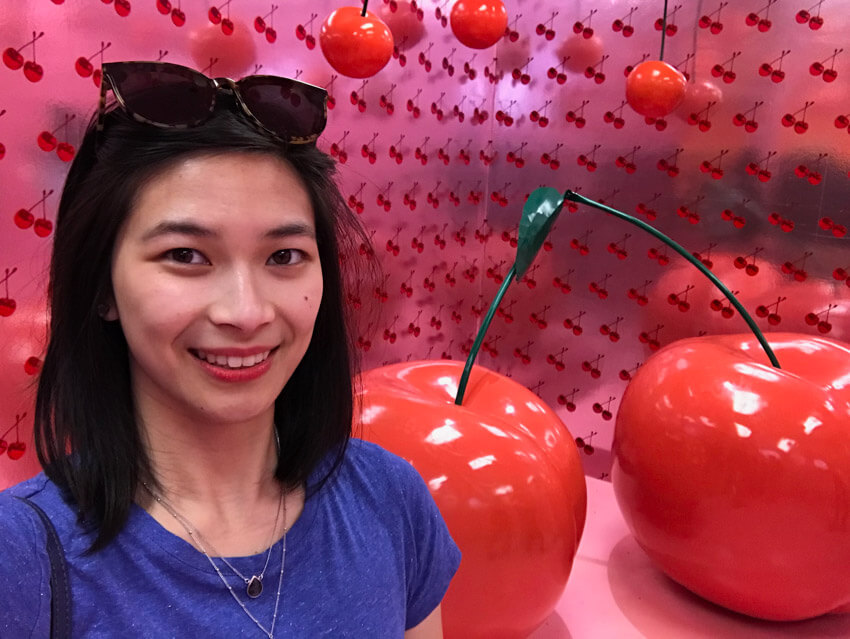A woman with short dark hair in a blue t-shirt. Behind her is a big sculpture of two giant cherries. She is in a room with pink wallpaper with a cherry pattern.