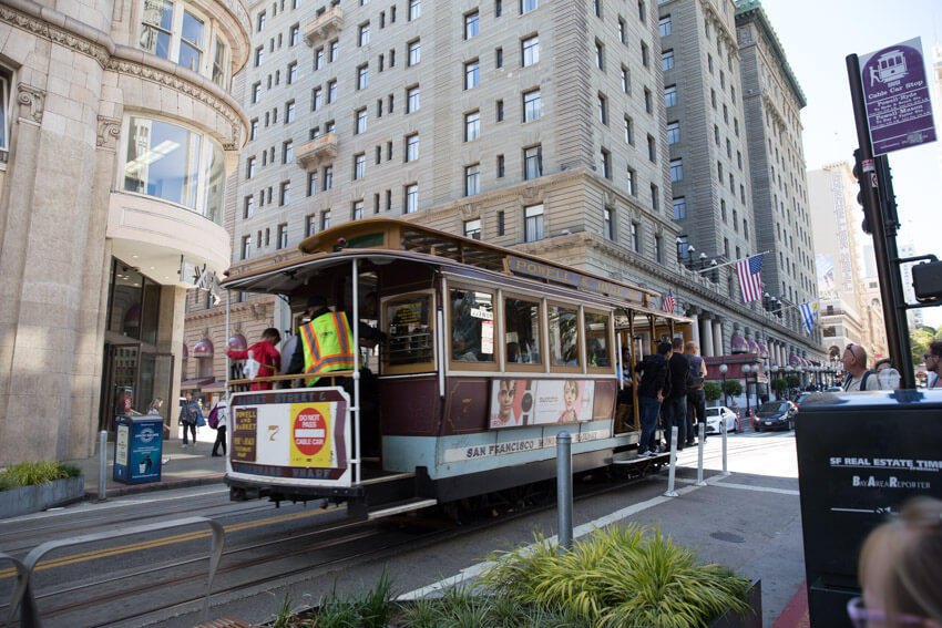 A cable car on a street with high-rise hotels in the background. Some people are standing and hanging onto the platforms at the side of the cable car.