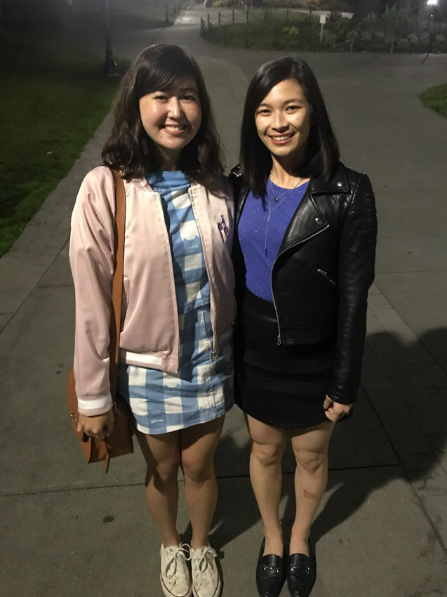 Two women joined at the hip, both have shoulder-length dark hair. One woman is wearing a blue and white dress with a pink bomber jacket; the other is wearing a blue shirt and a black skirt with a black leather jacket. It is evening and the women are standing in a park with a lamppost just behind them.