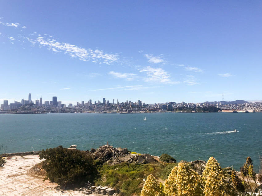A view from an island cliff face to the sea and the city of San Francisco opposite. There are a couple of boats on the water, leaving small trails of wake.