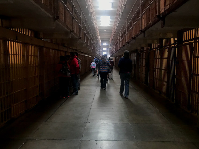 A corridor of jail cells with barred entrances. A bit of light flows in from the ceiling.