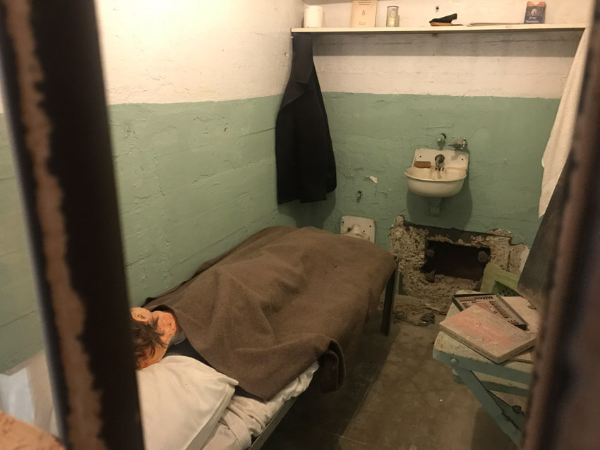 A jail cell with a model of a human head in the bed. In the wall is a small hole.
