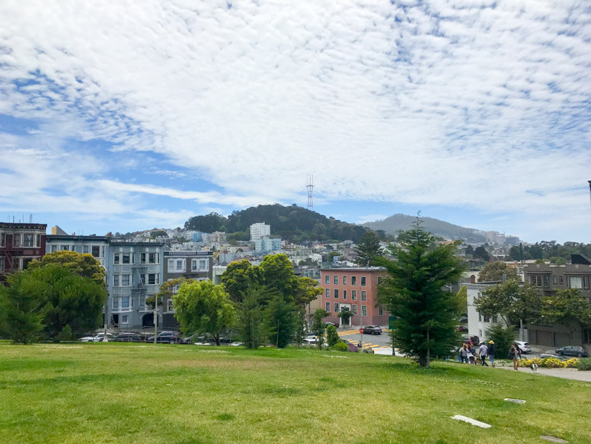 Part of the city of San Francisco, seen from the top of a green, grassy hill. In the distance, a lot of trees on a hill can be seen, as well as a communications tower. The sky is blue but filled with many scattered white clouds.