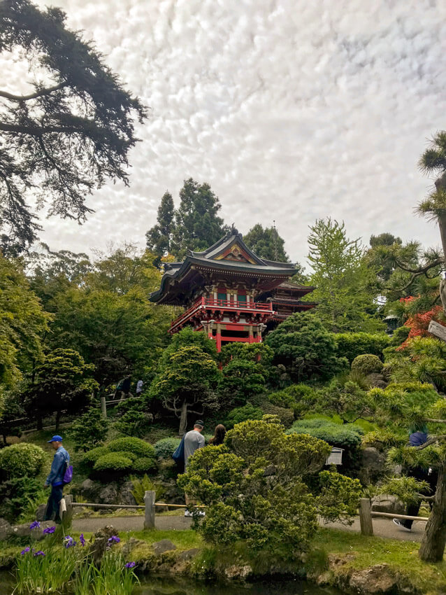 A Japanese garden full of green trees, with a Japanese-style temple house in the middle of the frame. The sky is full of white-grey clouds.
