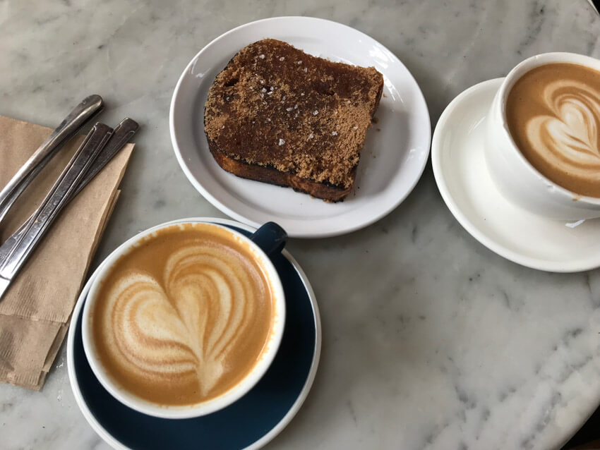 Two cups of coffee with heart-shaped coffee art, sitting on a marble surface, along with another plate of thick toast.