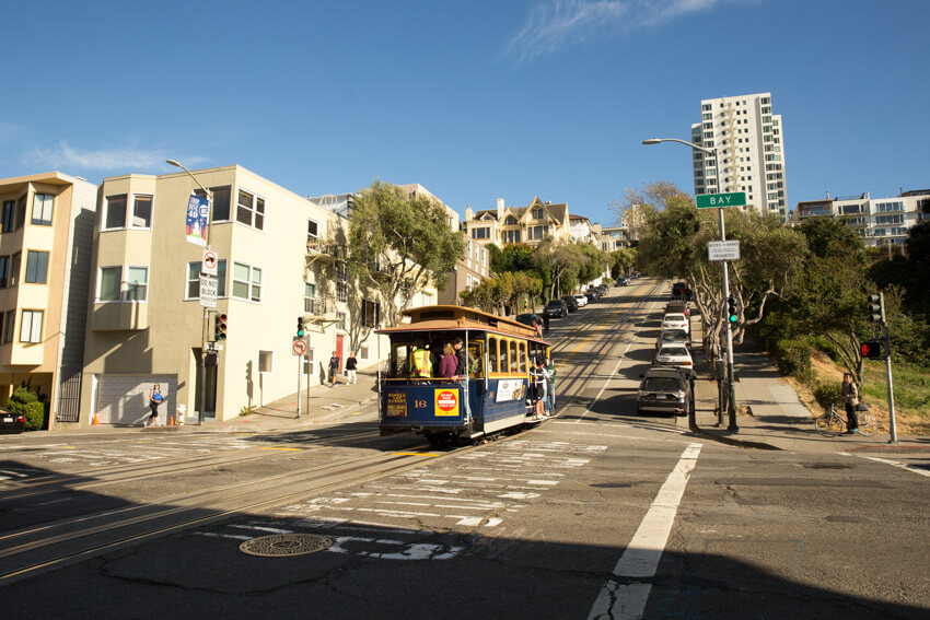 A cable car at an intersection, at the foot of a hill. It’s afternoon and the foreground has a fair bit of shade while the sky is blue.