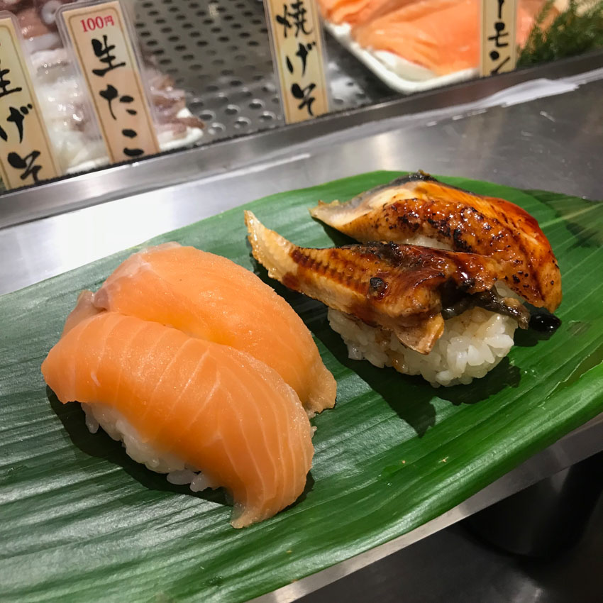 Four pieces of nigiri sushi on a large green leaf. Two are raw salmon, orange in colour, and the other two are grilled eel.