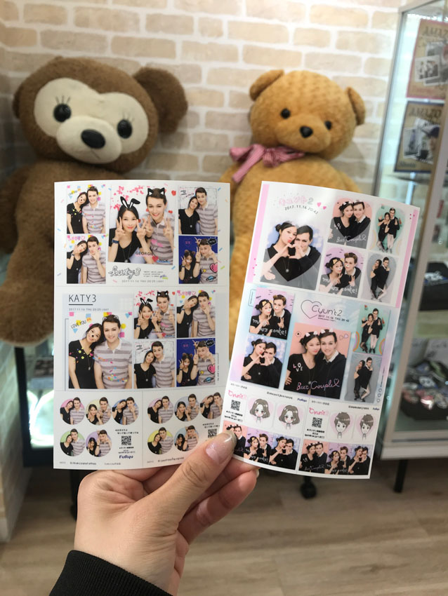 A hand holding two sheets with two sets of photos with a young couple (a boy and girl) smiling. Some of the photos are decorated with animal ears and colourful backgrounds. In the background is a big brown teddy bear and a big golden brown teddy bear.