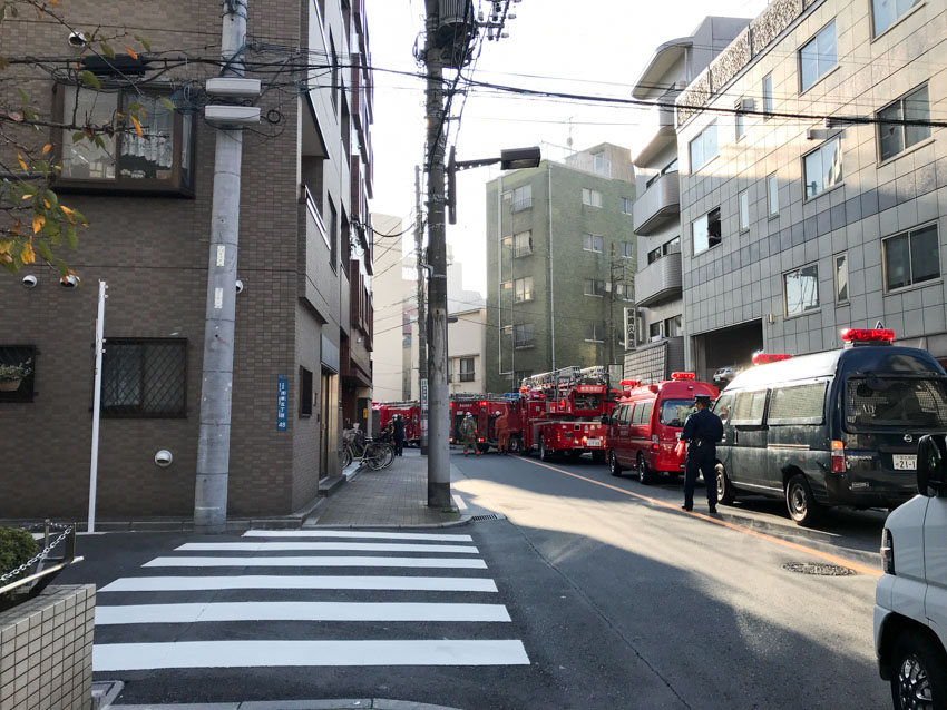 A street view with a street leading to the left, and a pedestrian crossing. There are a lot of red fire trucks in a line up the street. A police officer can be seen standing on the road.