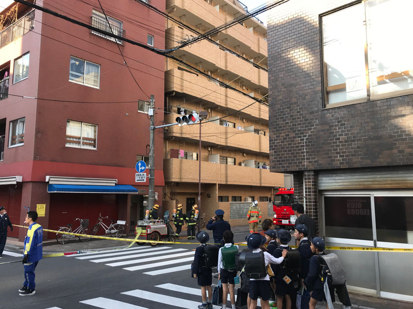 Apartment buildings at a residential intersection with access to one street blocked off by yellow tape. A small group of schoolkids are in front of the tape. Firefighters stand behind the tape, looking up at the second storey of an apartment building. The front of fire truck can be seen inside the street that is blocked off.