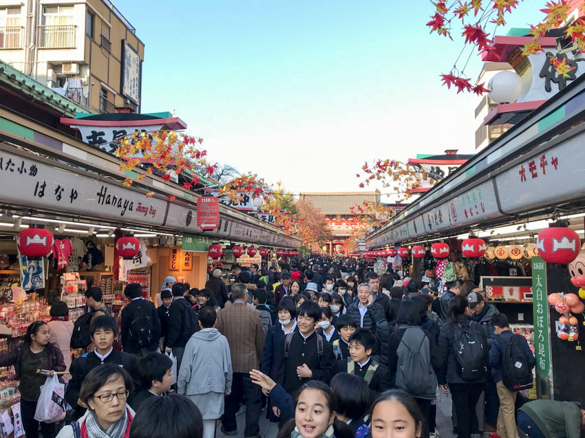 A street full of schoolchildren and people, with shops lining the sides. The shops have Japanese signage above them.
