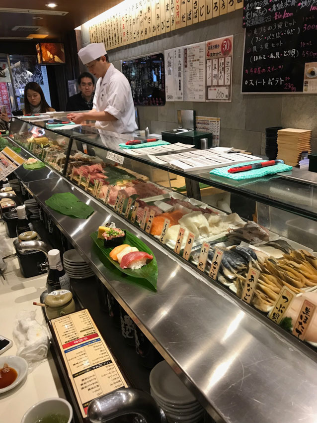 An angled side view of a standing sushi bar. There is a chef behind the counter dressed in white, some sheets of paper and pens on the glass counter, and various raw fish in the glass counter display. There are some condiments and sauces on the opposite side of the glass counter.