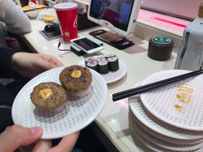 Close up view of someone holding a small white plate with a subtle textured checkered pattern, with two pieces of nigiri sushi, topped with small beef patties and yellow sauce. In the background is a sushi bar setting, with a computer screen, another plate of six maki rolls, and a small stack of empty plates in the same pattern.